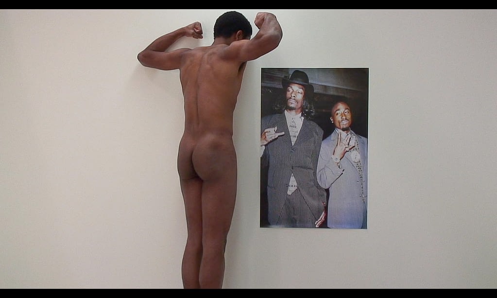 A slender black man stands with his back to the camera flexing before a poster depicting Tupac Shakur and Snoop Dogg