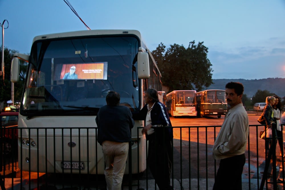 Man looks directly at camera whilie another man watches a video projected on the window of a bus