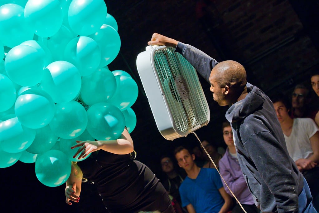A man holds a boox fan near a woman whose head is obscured by a large collection of turquoise balloons