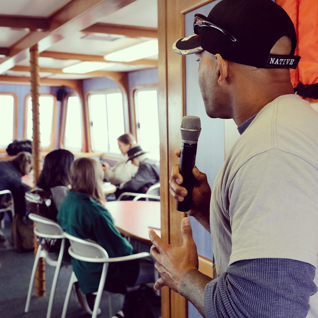 A brown man with a baseball cap with the text "Native" speaks into a microphone before people on a boat.