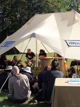 A large group gathers under a shade tent for a discussion.The tent has a banner reading "Indigenous"