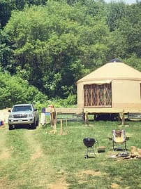 A beige yurt, campfire and a truck in a wooded clearing