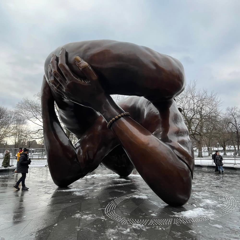 A few people in a snow-dotted plaza view a monumental bronze sculpture in a knotted embrace under a cloudy winter sky. 