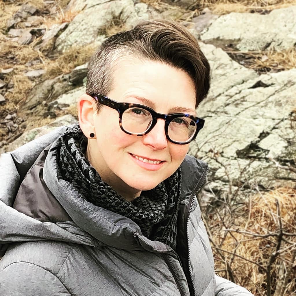 Image of a pale-skinned person with glasses and a side-shave pompadour hair cut wearing a gray parka and standing in front of rocks.