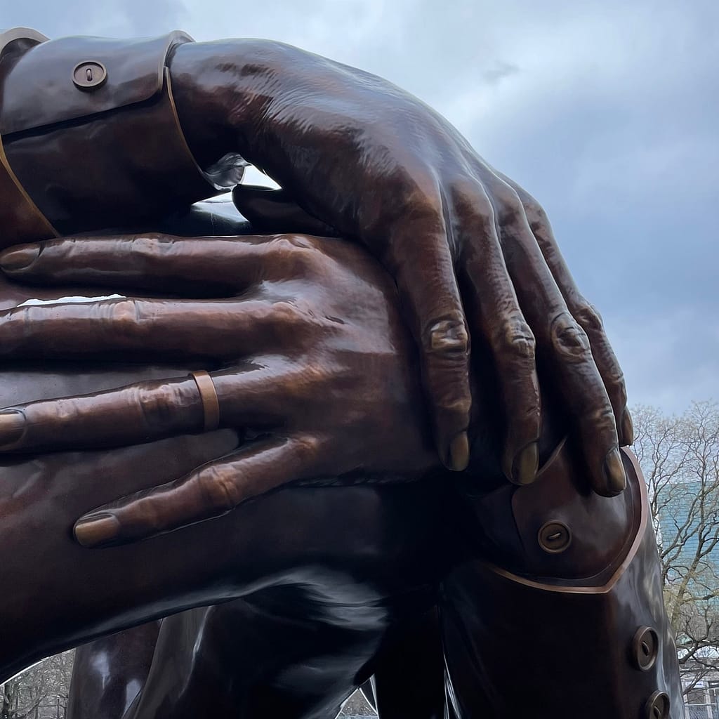 Monumental bronze hands (one with a wedding ring) clasp against a stormy winter sky.