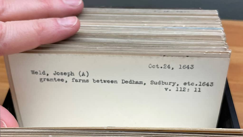 Pale fingers opens reference cards in a card catalog. The card reads "Weld, Joseph A. grantee, farms between Dedham,  Sudbury, etc. 1643"
