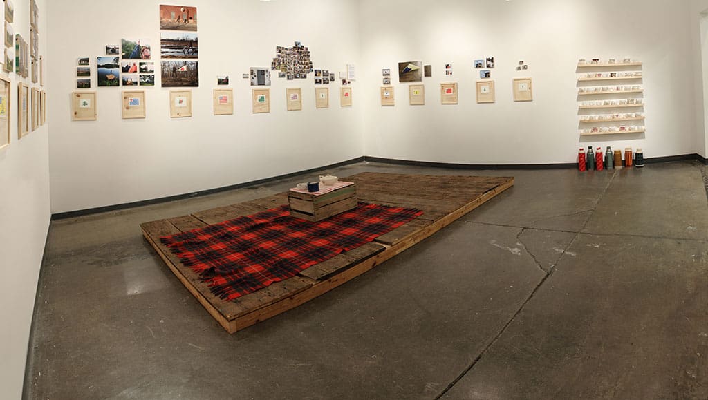 Gallery installation with small objectson wall and platform with plaid blanket on the floor