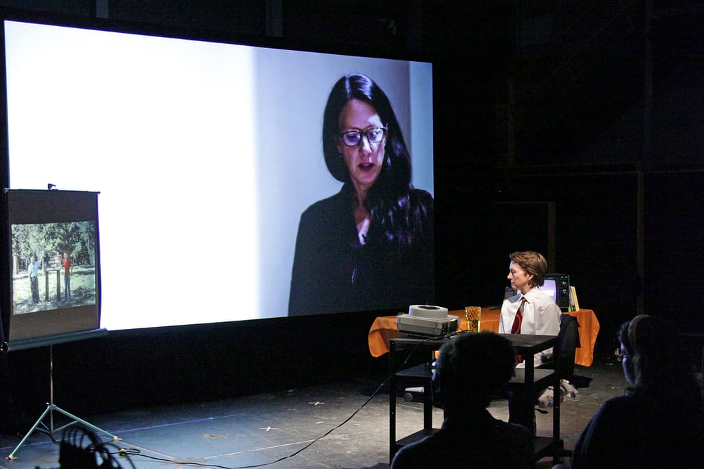 The artist Sarah Kanouse sits to the right side of an image of a performance space and gazes at a 35mm slide projection of a family image. A large-scale projection showing another White-presenting woman giving a lecture is shown in the background.