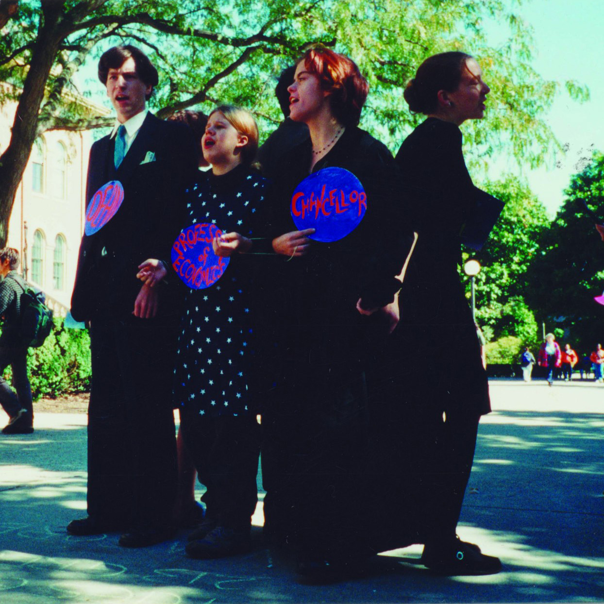 Five people dressed in black clothing with hand-painted labels describing positions in a university stand in a circle during a street theater performance.