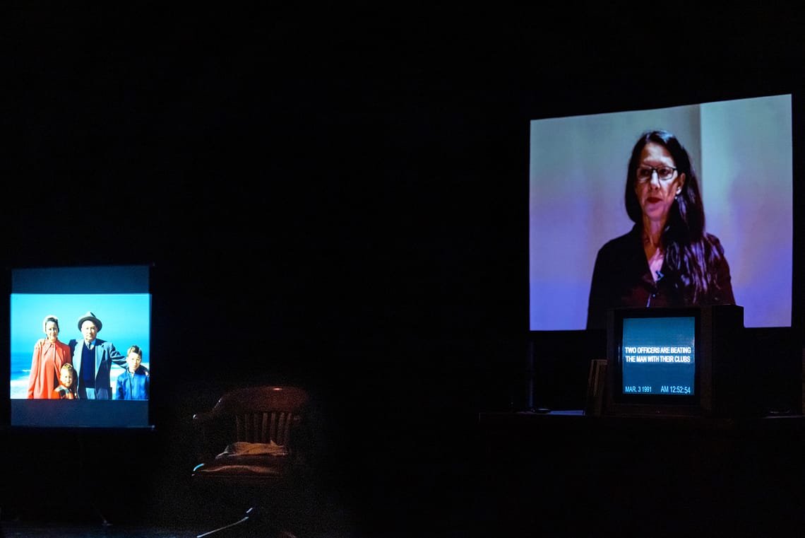 Three screens on a stage show various images: a projected image of a white, 1950s family, a white, female-presenting person at a podium, and a TV screen with a transcript of police conduct from the Rodney King beating video.