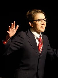 The artist performing in a blue men's suit, white shirt, red tie and midcentury glasses. Her right hand is raised.