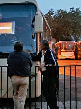 Two male-presenting people face away from the camera watching a video projected on the front windshield of a bus.