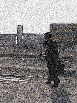 Highly pixelated and noisy image of a female presenting figure walking throug an industrial landscape with a case and an antenna.