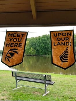A picnic shelter frames a view of a bench looking out on a river. Two tan and green cloth banners hang from the shelter reading "Meet you here" and "Upon our lands"