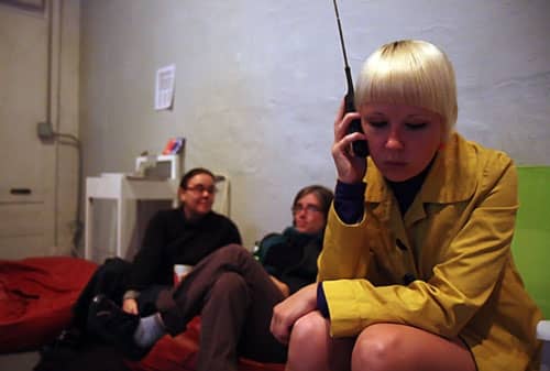 A female-presenting person with light skin and bleached hair listens to a handheld radio in the foreground while two other light-skinned people lounge in soft focus in the background.