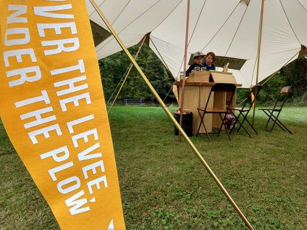 Yellow pennant reading "Over the Levee Under the Ploy" in foreground with wooden table on grass beneath tent in background with children.