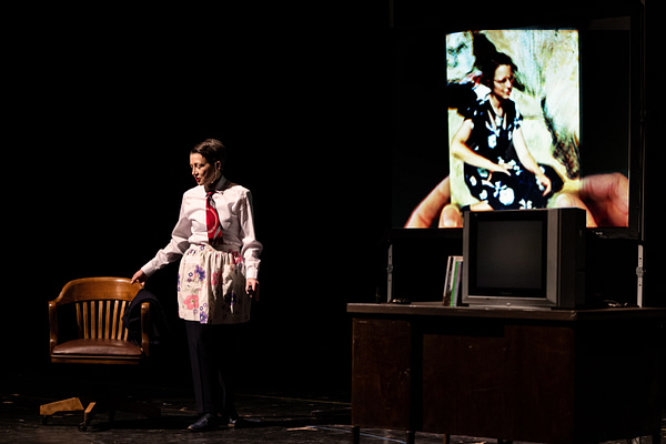 The artist Sarah Kanouse reaches for a chair while wearing a red tie and floral apron during a scene from "My Electric Genealogy." A projection of hands holding a photo of the artist's grandmother displays on stage.