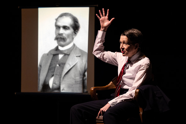 Artist Sarah Kanouse raises her hand with an open palm beside a projected image of an African American man in a 19th century suit in a moment from the performance "My Electric Genealogy"