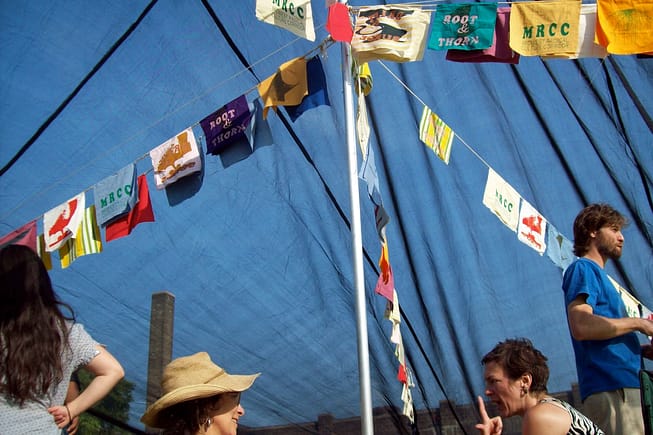Low angled image of interior of sun shade hung with multicolored, handmade pennants. Light-skinned people converse on the edges of the fram.