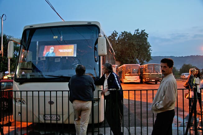 Two male-presenting people face away from the camera watching a video projected on the front windshield of a bus. Another man walking by gazes directly into the camera.