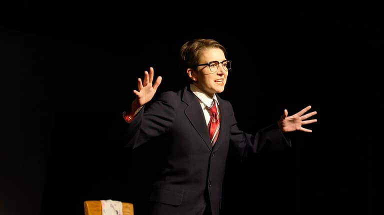 Alt: Light skinned, female-presenting figure in a men's suit, glasses, and a red tie stands beside in a dark space beside a wooden chair, both hands raised in an animated expression.